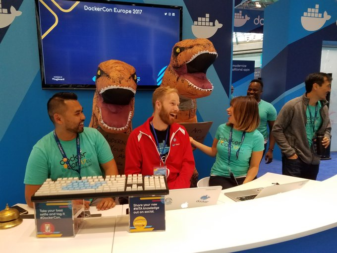 Photo of person in brown dinosaur suit behind seated people at DockerCon booth.