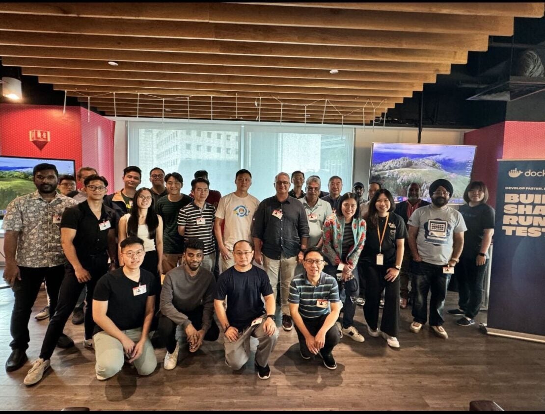 Photo showing a group of people sitting and standing in front of a large window at a Docker DevTools event.