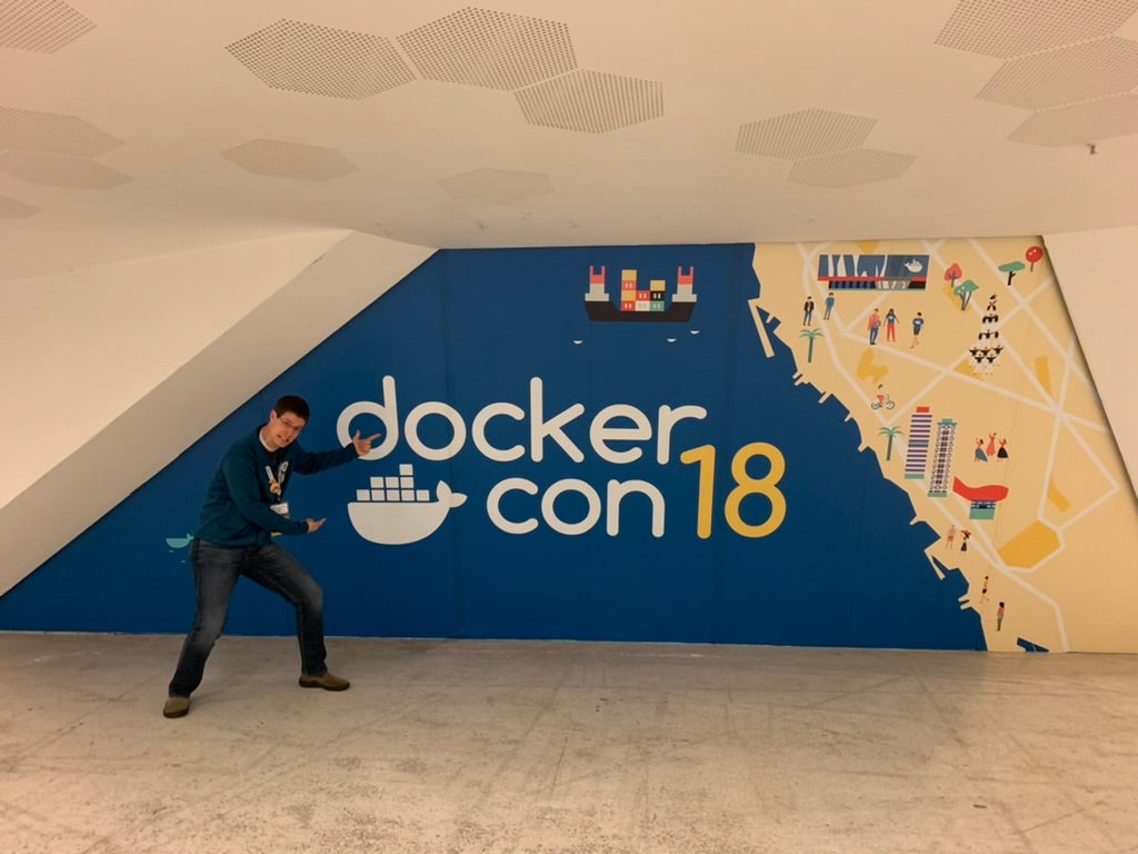 メイン基調講演室の外に設置されたDockerCon EU 2018 の看板(バルセロナ)を指差す黒髪の男性を示すマイケル・アーウィンの写真。